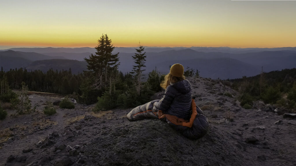 A backpacker sitting on a rock in the REI Magma 30 sleeping bag at sunset