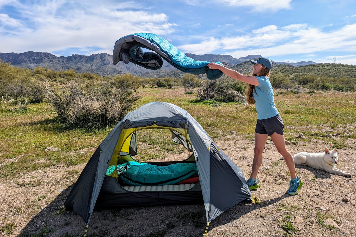 A woman unfurling the Mountain Hardwear Phantom 0 backpacking sleeping bag over a tent with a beautiful mountain view behind her.