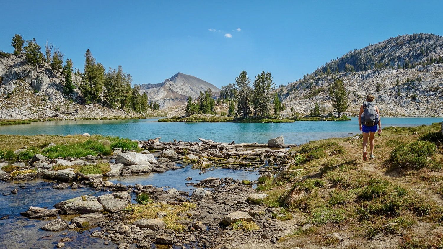 A backpacker hikes by the edge of a turquoise blue lake in the Wallowa mountains with a beautiful island in the middle