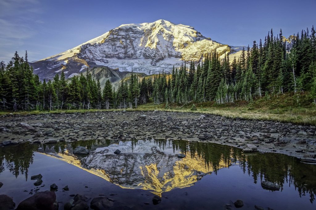 A snow-capped mountain is reflected in an alpine lake, with trees surrounding the lake.