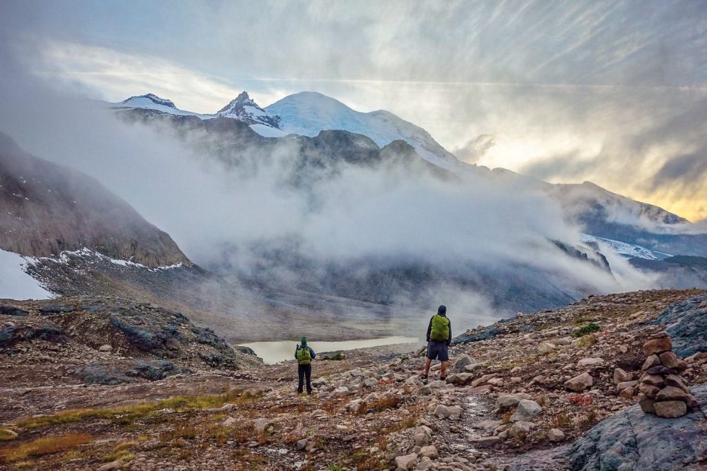 Hikers walk across a scree field with a clouded mountain in the background.