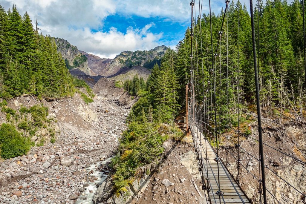 A suspension bridge crosses a river along the Wonderland Trail, with views of distant peaks in the background.