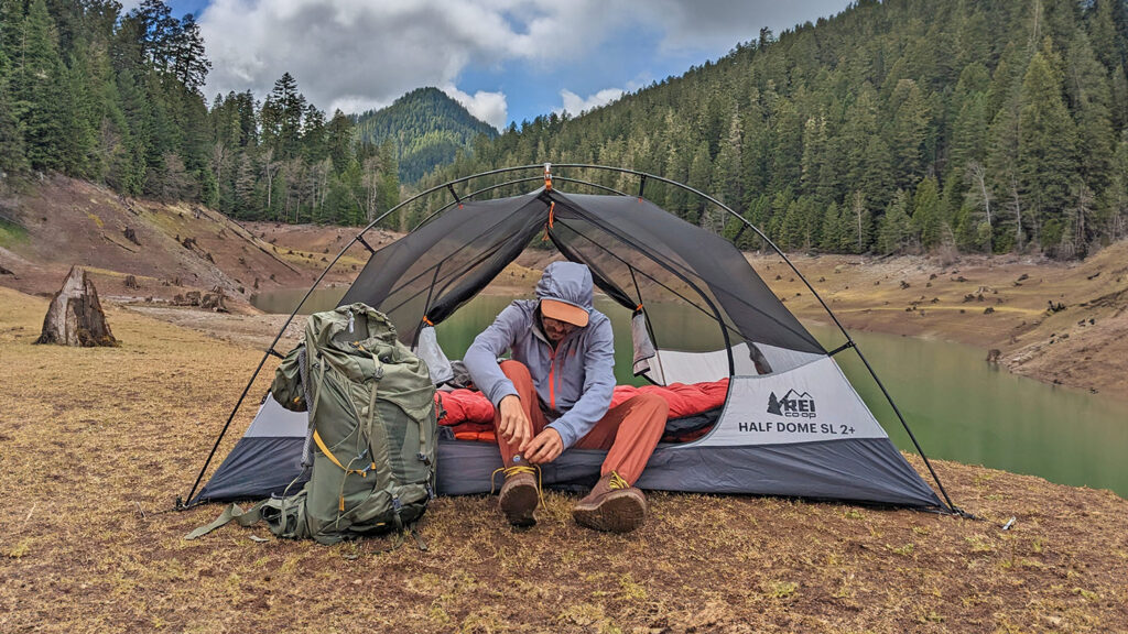 A hiker sitting in the doorway of the REI Half Dome SL 2+ backpacking tent with a tree-lined lake in the background