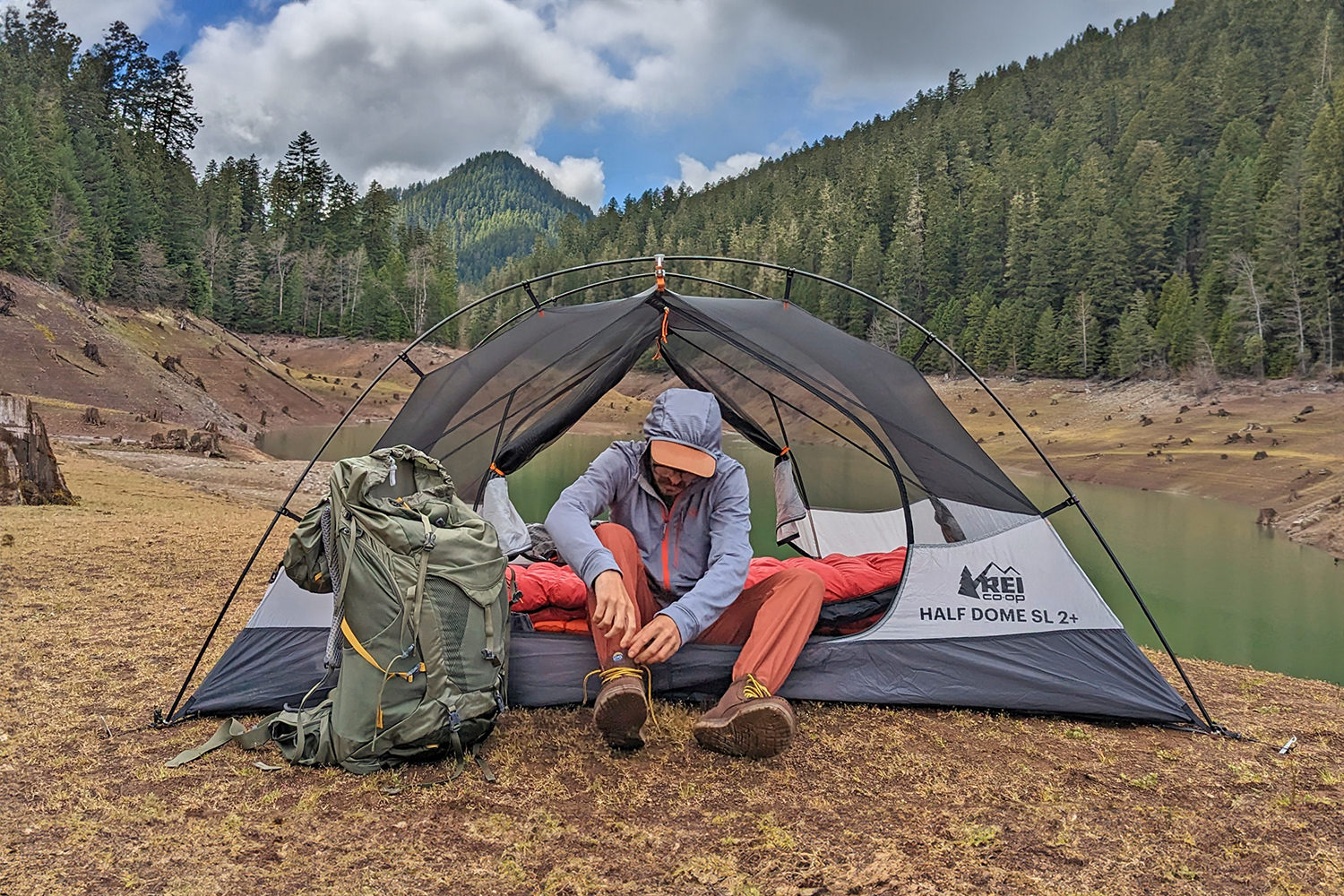 A hiker sitting in the doorway of the REI Half Dome SL 2+ backpacking tent with a tree-lined lake in the background