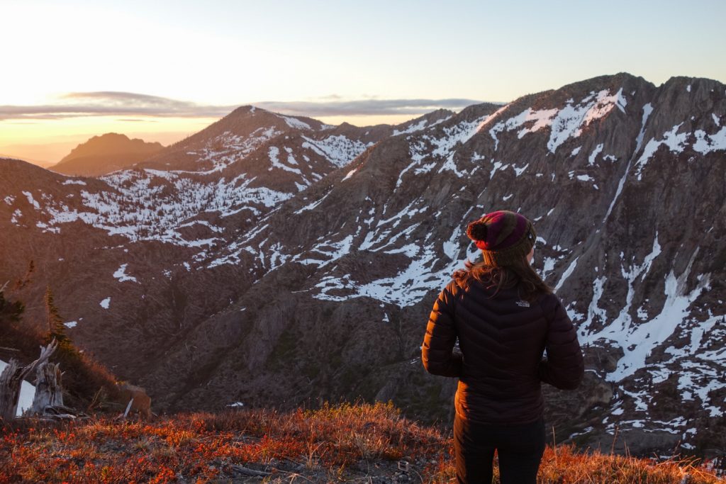 A hiker looks out as the sun sets over snow-capped peaks.