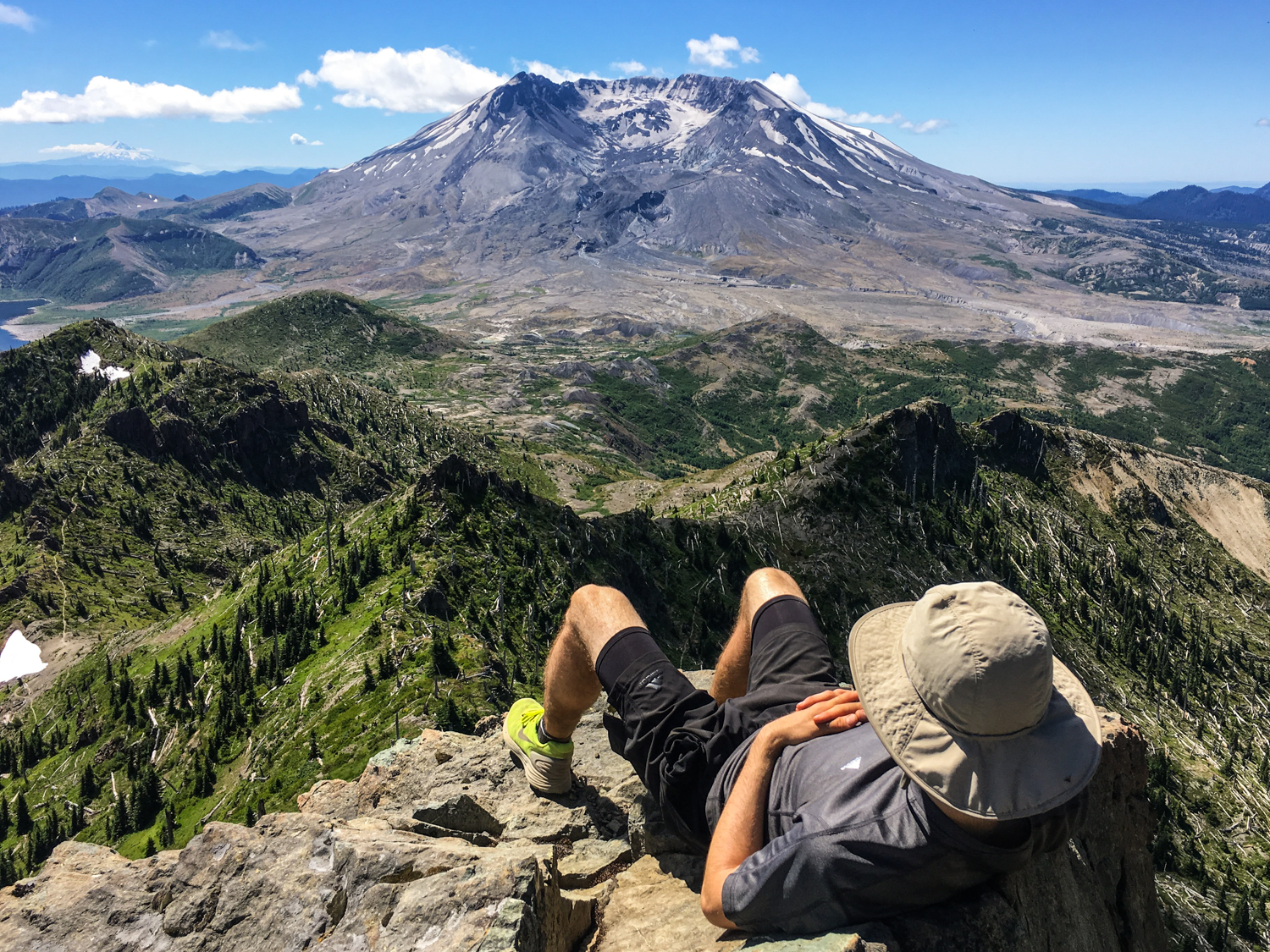 A hiker lies on a rock overlooking a dense forest with a large peak in the distance.