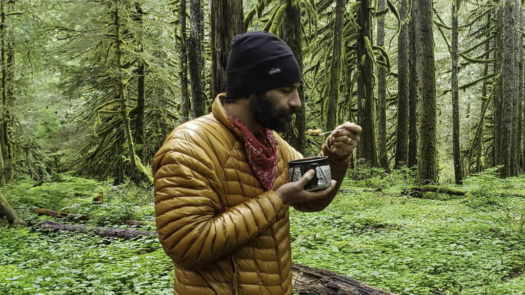 A hiker eating from a titanium backpacking pot in a lush, green forest