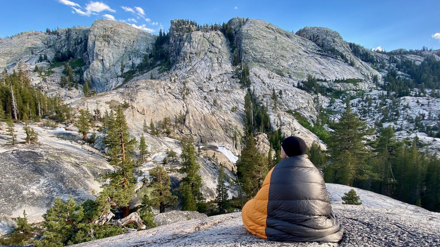 A hiker sitting on a ridge in the SIerra wrapped up in the Enlightened Equipment Revelation Quilt