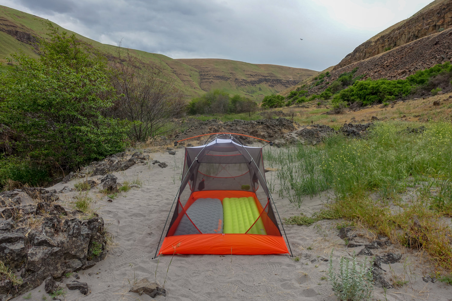 A tent from its narrow side framed in a sandy wash between two grassy hills.