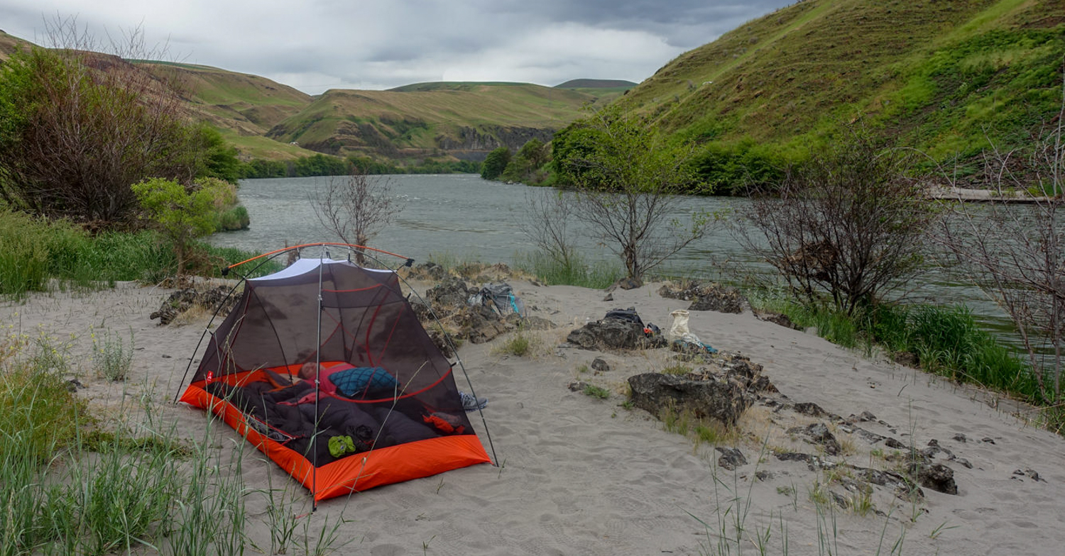 A person sleeps in a tent with no fly on a sandy wash with a lake nearby.