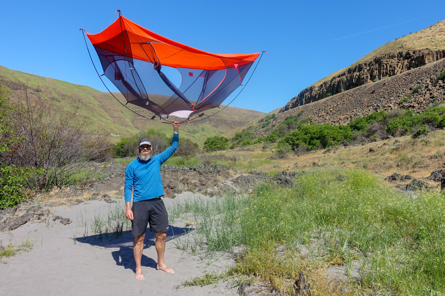 A tall, bearded man holds a tent high above his head with one arm in a sandy, grassy valley.