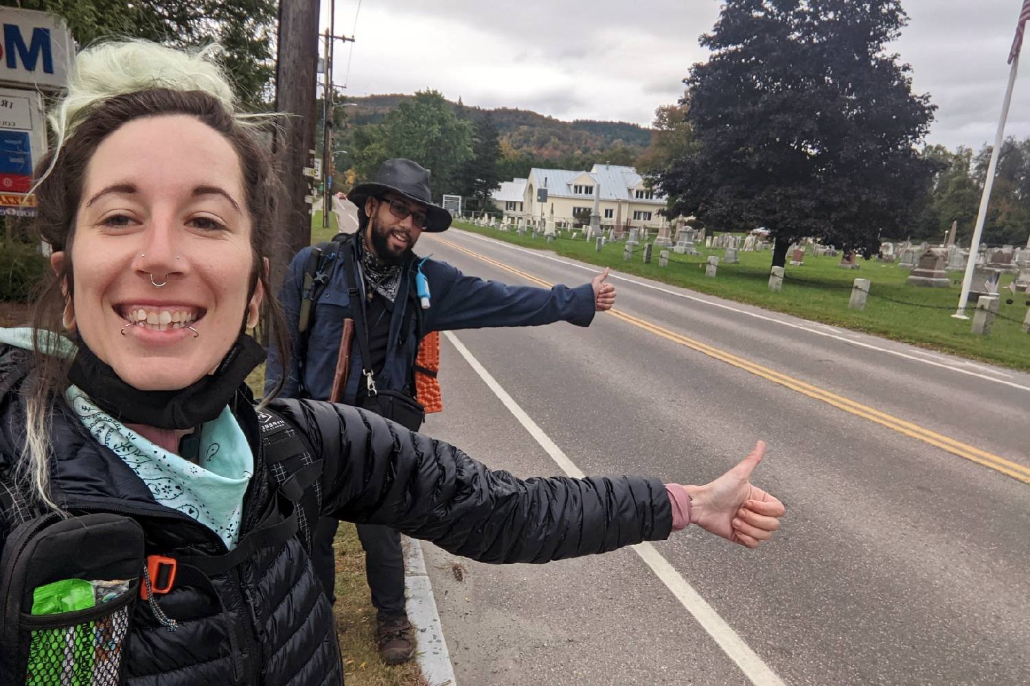 Two hikers holding out their thumbs to hitchhike out of a town
