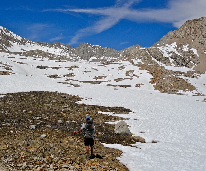 a man faces the camera with a big snow field behind him wearind trail runners