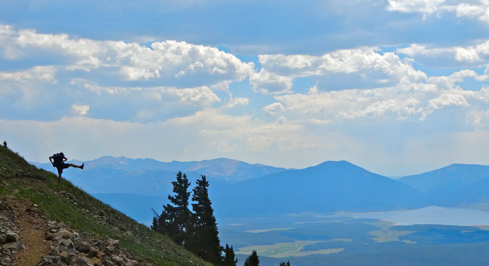 a silhouette of a man hiking a shoe into the horizon on a steep hill with mountains and a blue sky behind him
