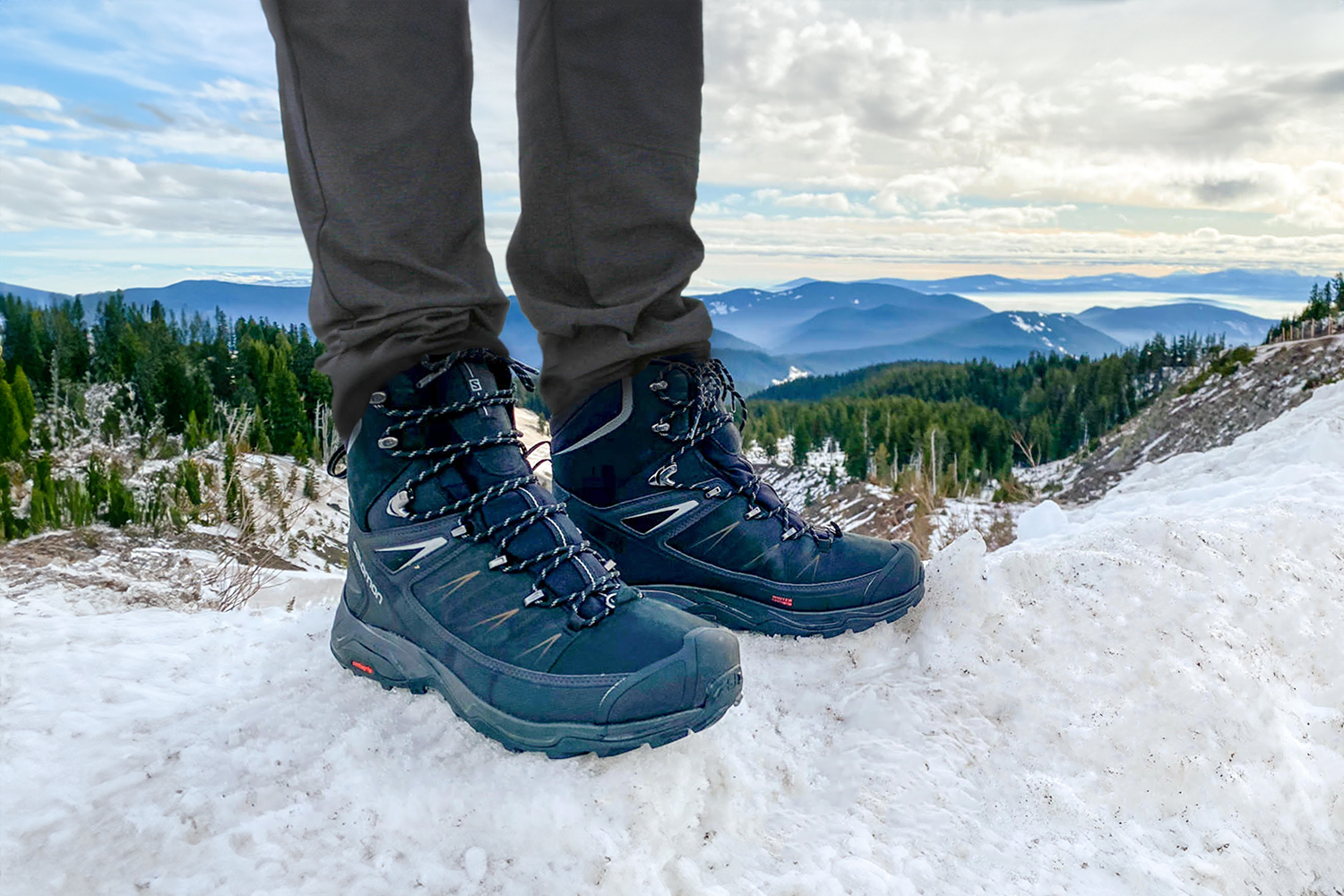 A hiker standing on a snowbank in the Salomon X Ultra Mid Winter boots with blue mountains on the horizon