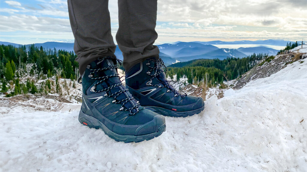 A hiker standing on a snowbank in the Salomon X Ultra Mid Winter boots with blue mountains on the horizon