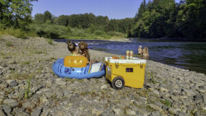 A woman and a puppy sitting in a float tube next to the Rovr Rollr 60 cooler at the river's edge