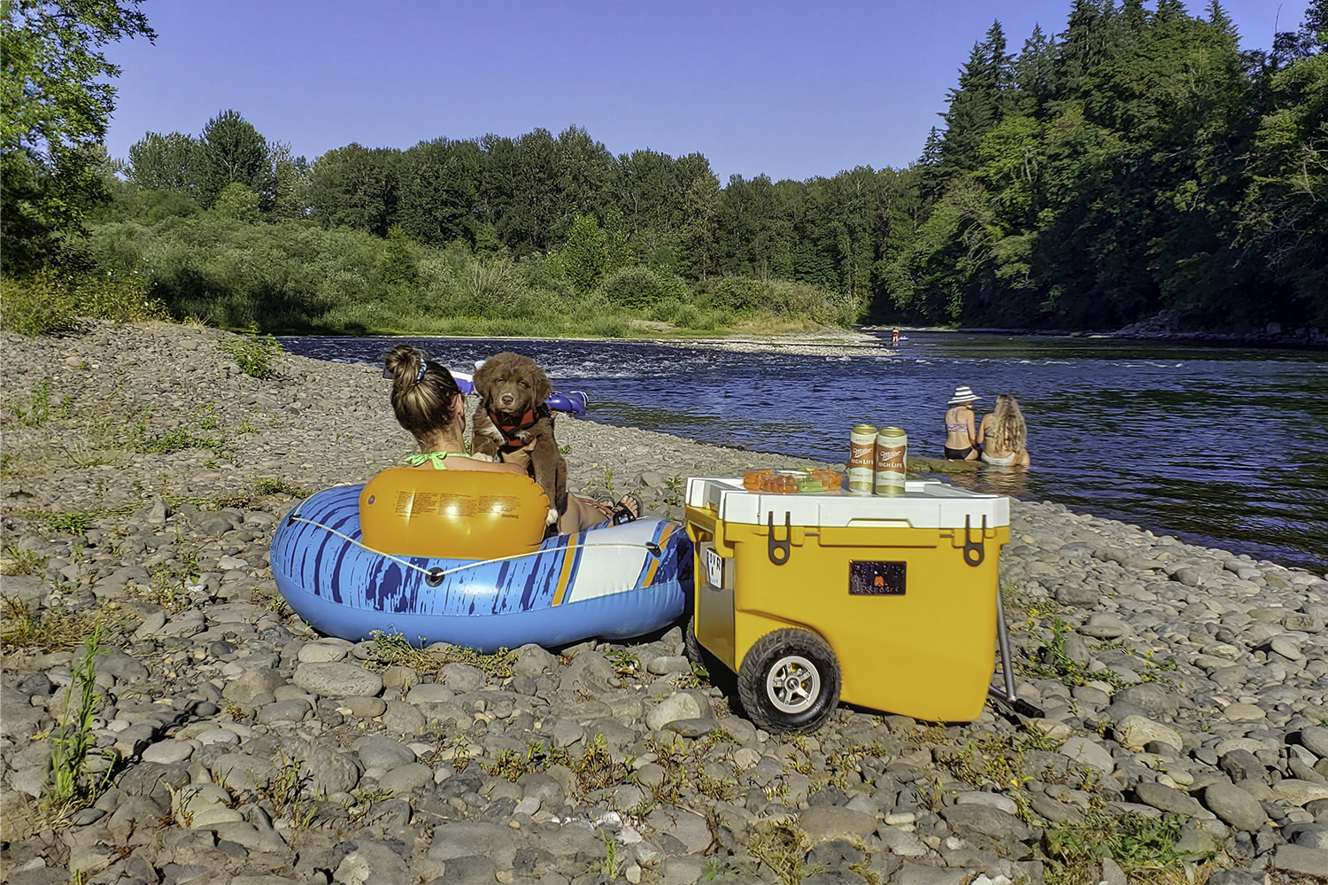 A woman and a puppy sitting in a float tube next to the Rovr Rollr 60 cooler at the river's edge