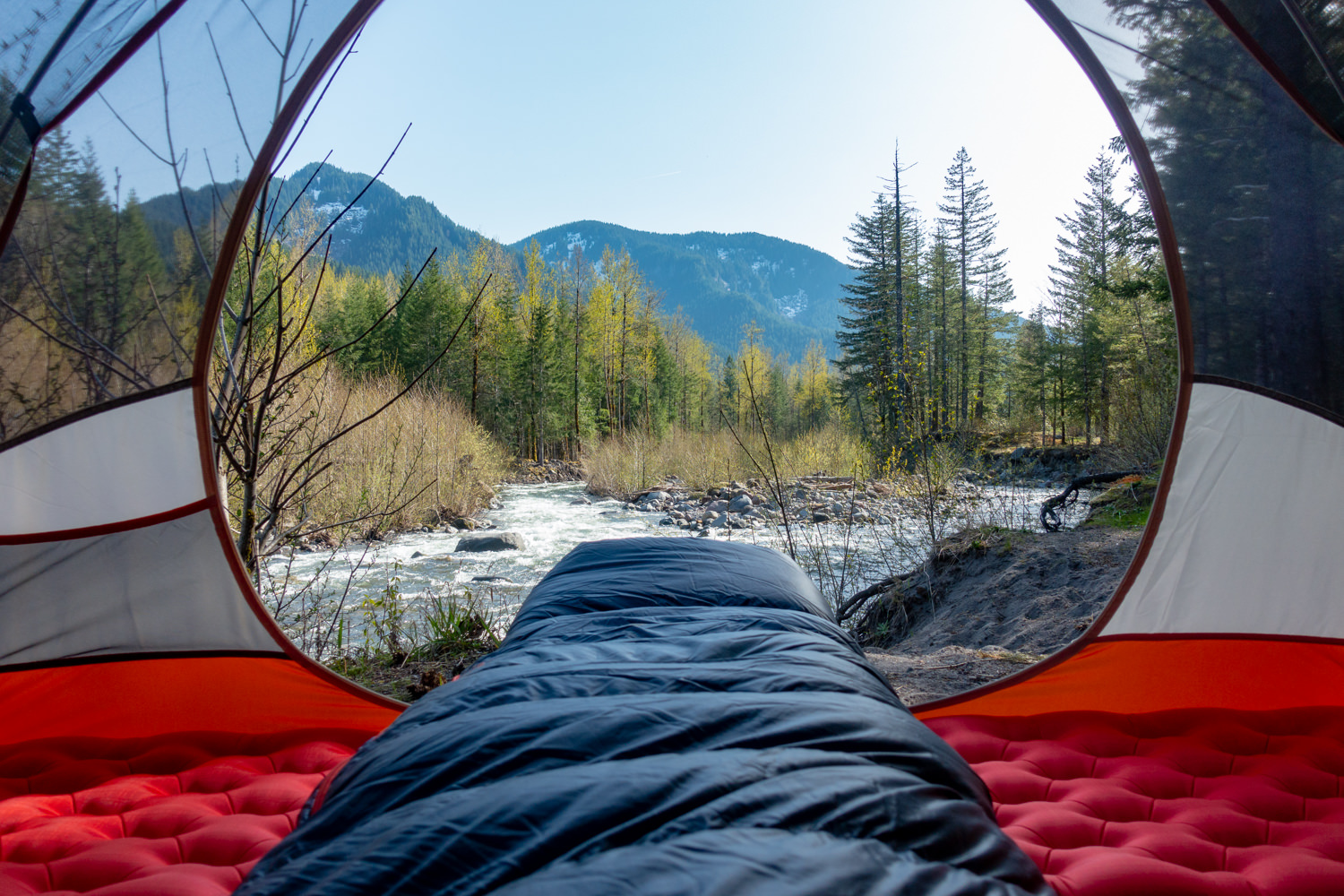 Looking out the doorway of a backpacking tent over the foot of a sleeping bag with a river and mountains in the background