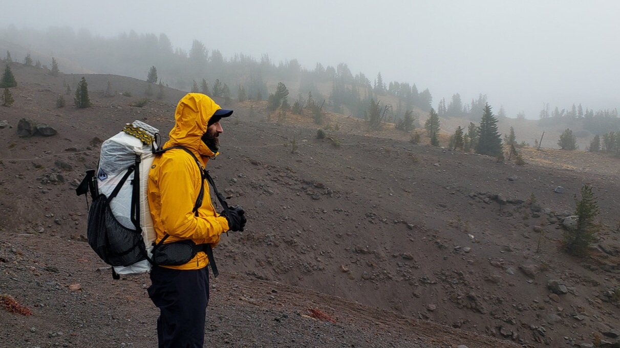 A backpacker in a rain jacket in front of a misty mountain ridge.