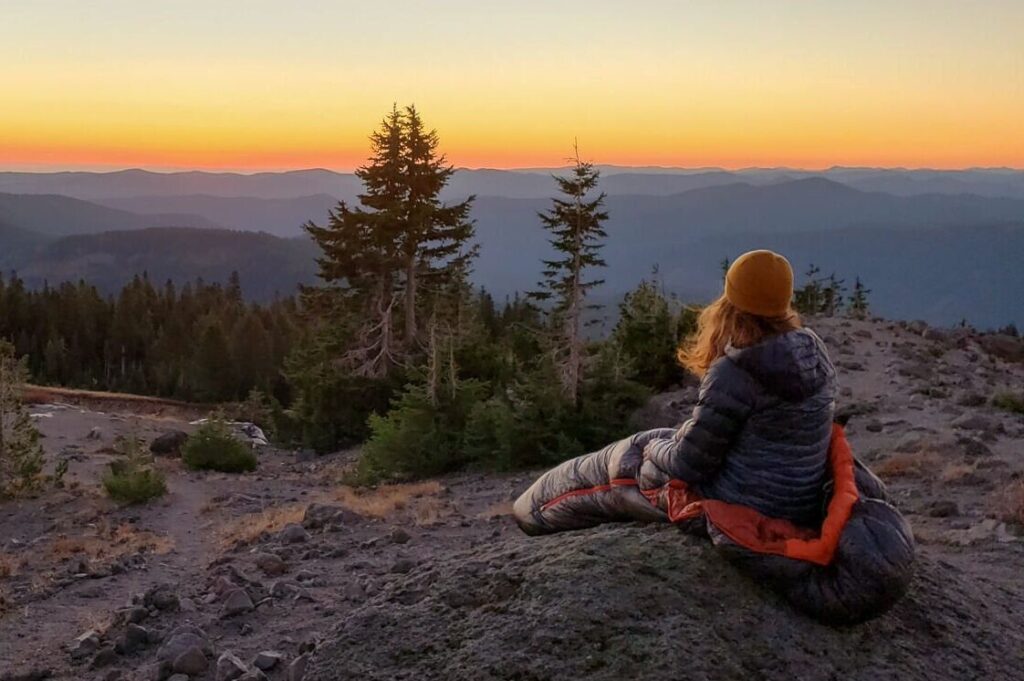 A backpacker sitting on a boulder in the REI Magma 15 sleeping bag while overlooking a mountain view at sunset