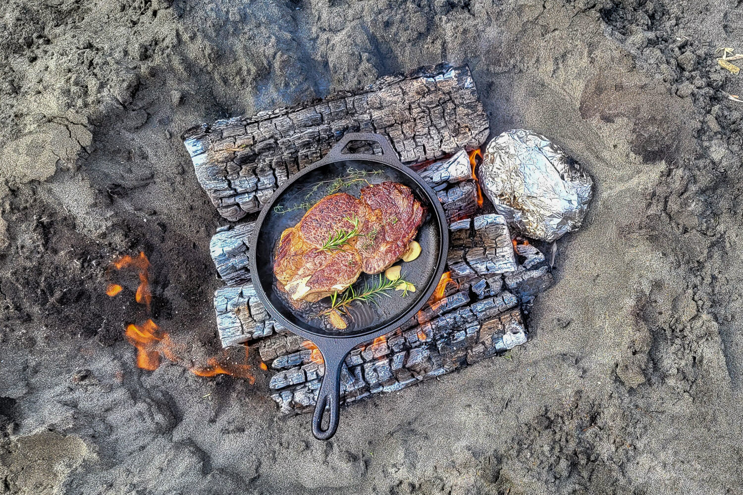 Using the Lodge Dutch Oven Combo Cooker to sear a ribeye steak over the fire - potatoes are wrapped in foil on the side