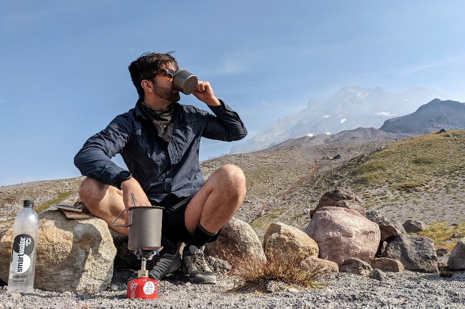 A hiker drinking from a cup with a mountain in the background - the Snow Peak Litemax stove is set up in front of them