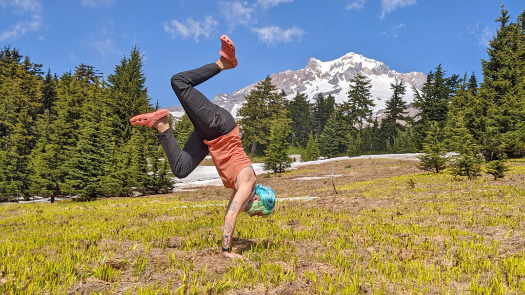 A backpacker doing a handstand in a mountain meadow in a pair of pink Crocs Classic Clogs