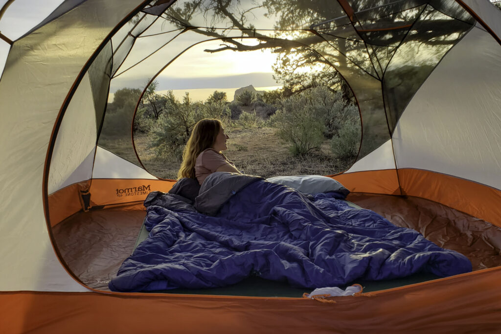 A woman snuggled up in the Exped MegaSleep 25/40 sleeping bag inside of a camping tent with a view of the high desert in the background