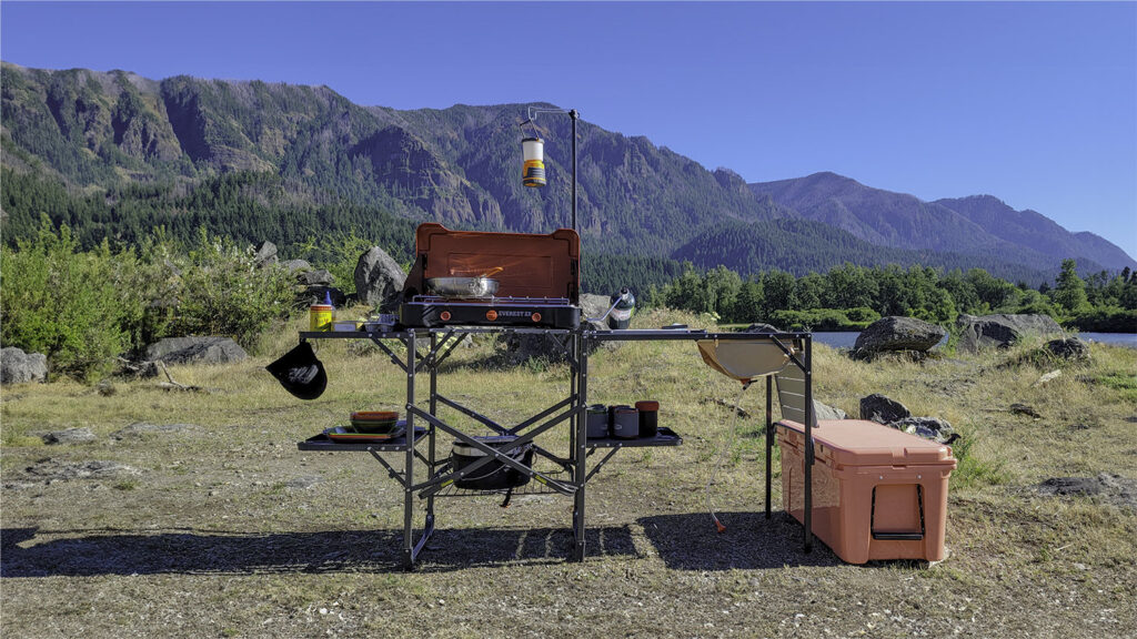 The GCI Outdoors Master Cook Station set up in a lakeside campsite with the Camp Chef Everest 2x stove, a cookset, a YETI Tundra Cooler, lantern, etc.