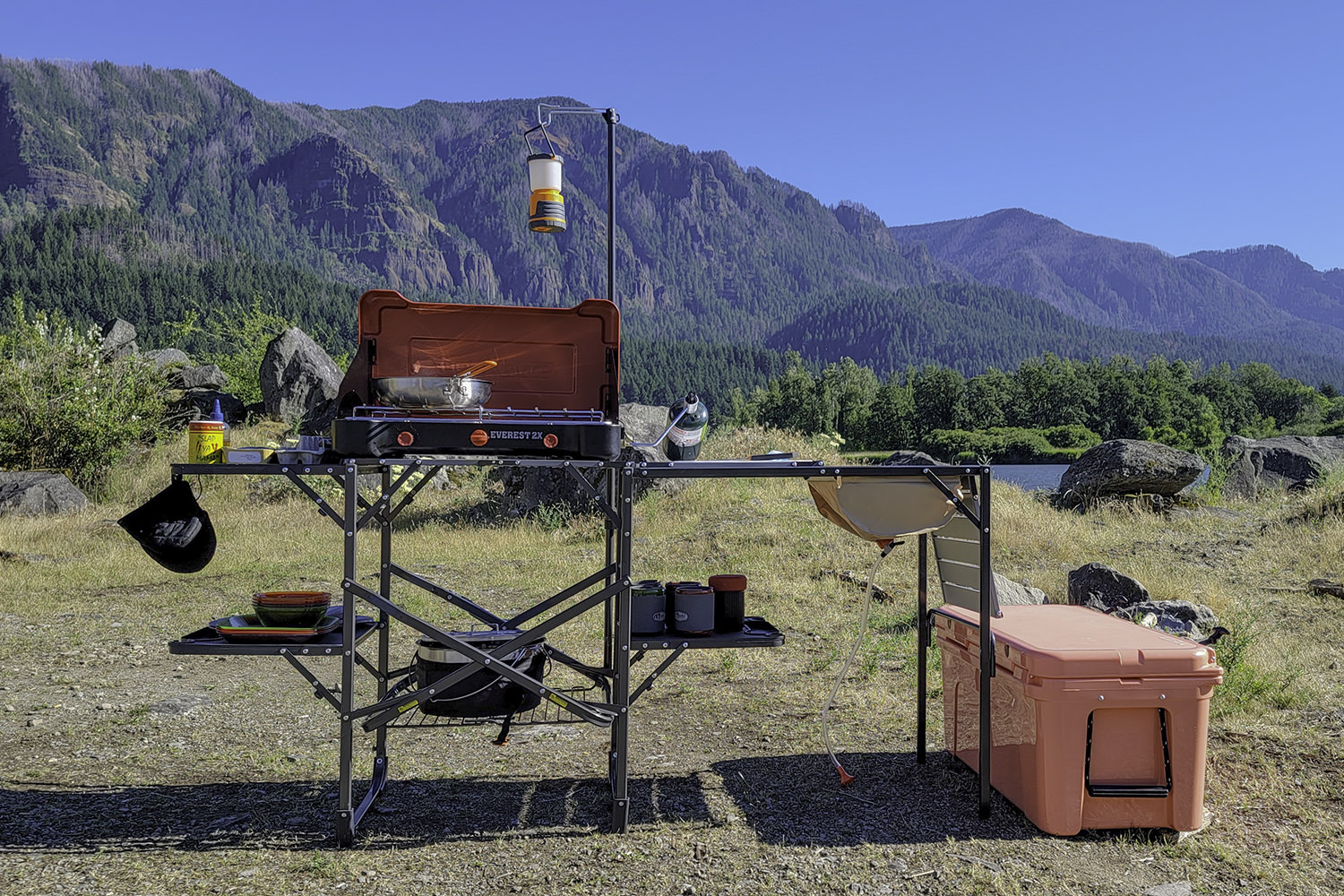 The GCI Outdoors Master Cook Station set up in a lakeside campsite with the Camp Chef Everest 2x stove, a cookset, a YETI Tundra Cooler, lantern, etc.