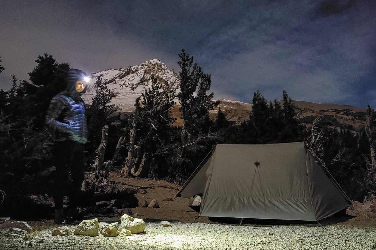 A hiker shining light from the Petzl Actik Core headlamp on a tent with a mountain in the background