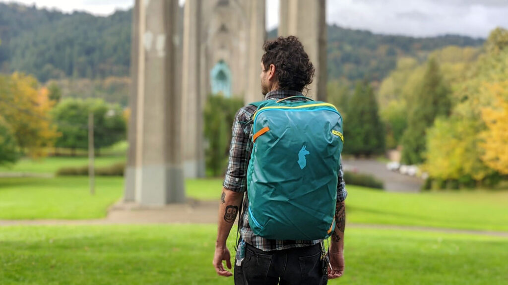 Back view of a guy wearing the Cotopaxi Allpa travel backpack in a green park with a bridge overhead
