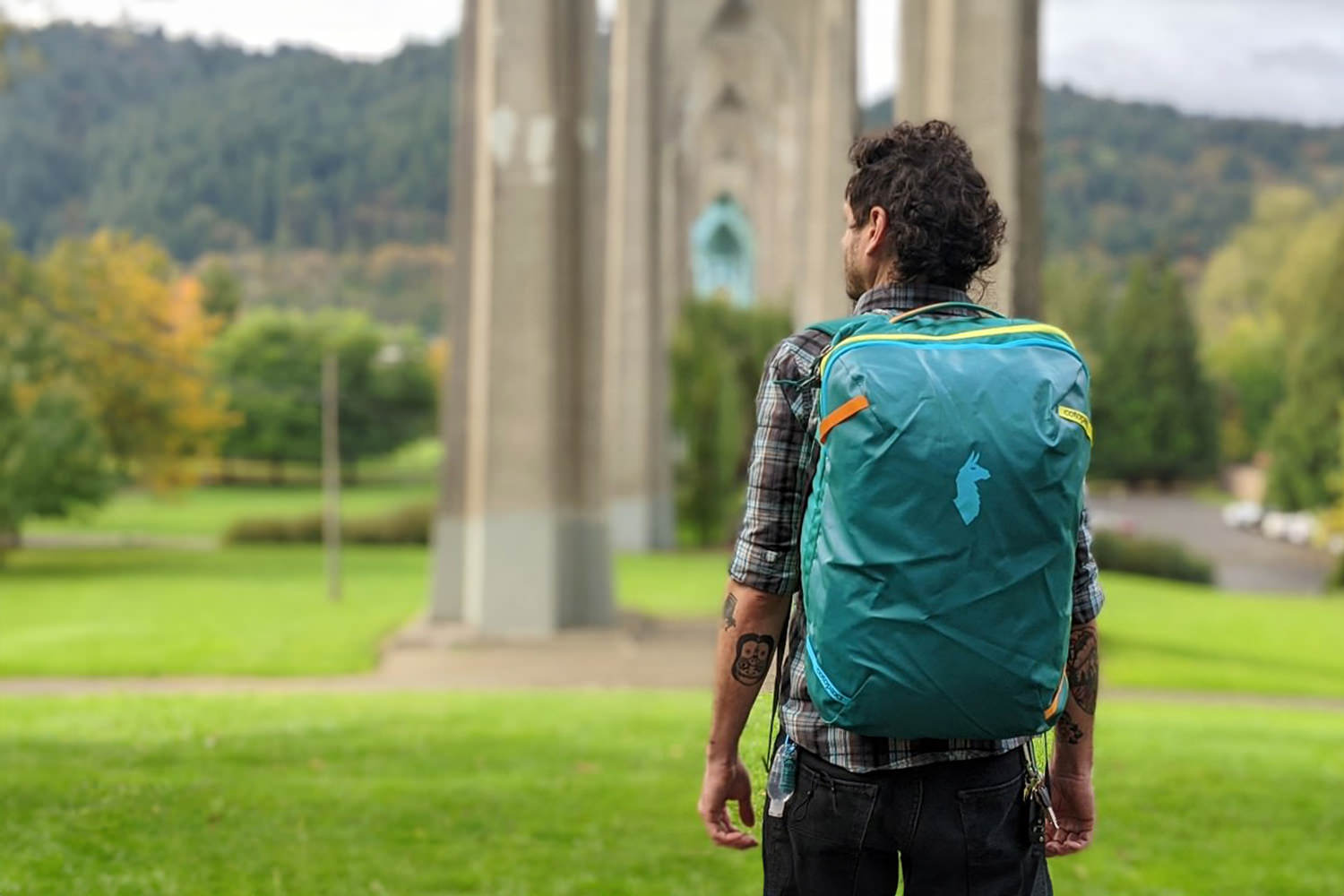 Back view of a guy wearing the Cotopaxi Allpa travel backpack in a green park with a bridge overhead