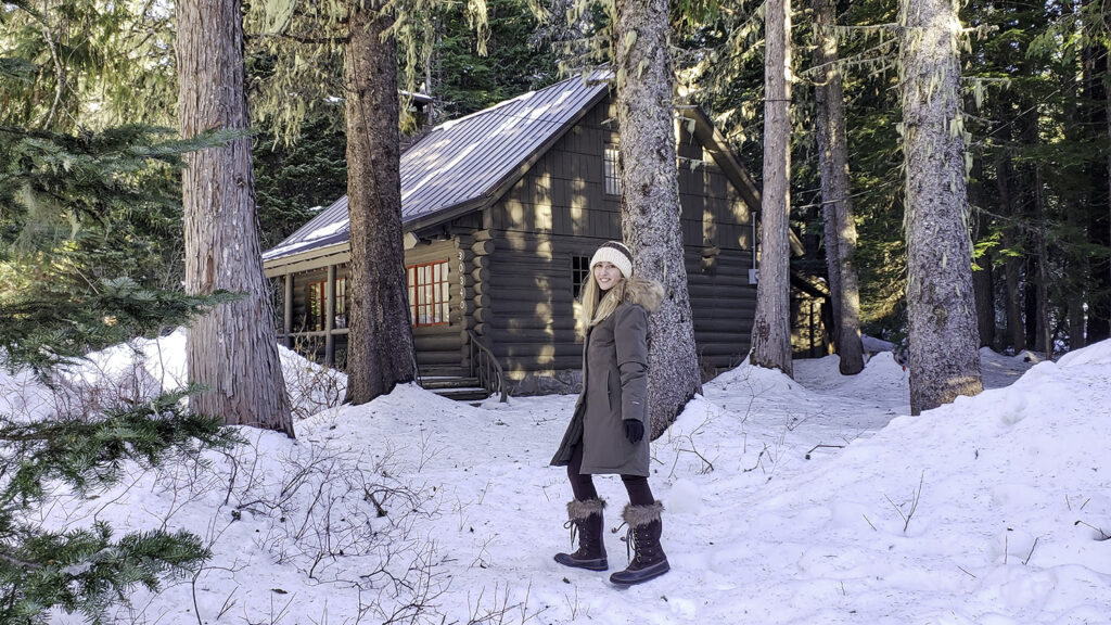 A woman standing in the snow in The North Face Arctic Parka outside of a rustic cabin in the woods