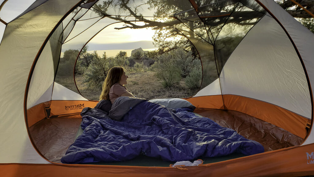 A woman snuggled up in the Exped MegaSleep 25/40 sleeping bag inside of a camping tent with a view of the high desert in the background