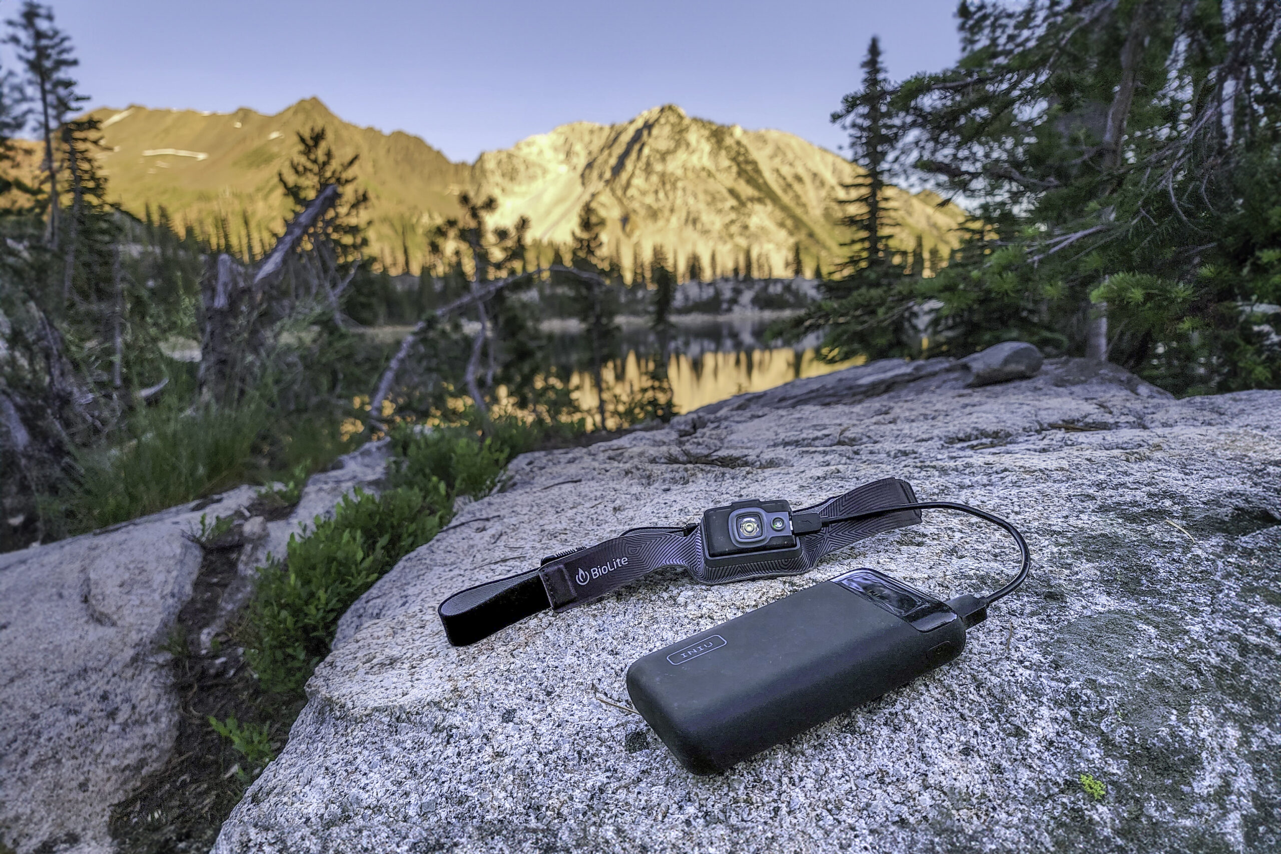 The Iniu 20000 PD power bank sitting on a granite slab while charging a rechargeable Biolite headlamp with the Wallowa Mountains at sunset in the background