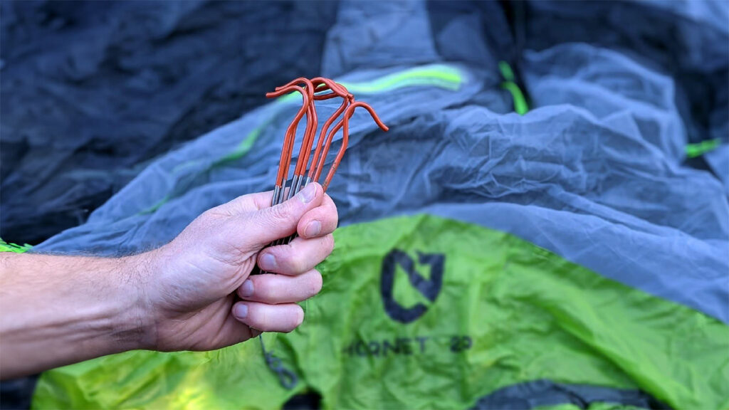 A backpacker's hand holding a set of Vargo Titanium Shepherds Hook tent stakes in his hand with a NEMO tent in the background