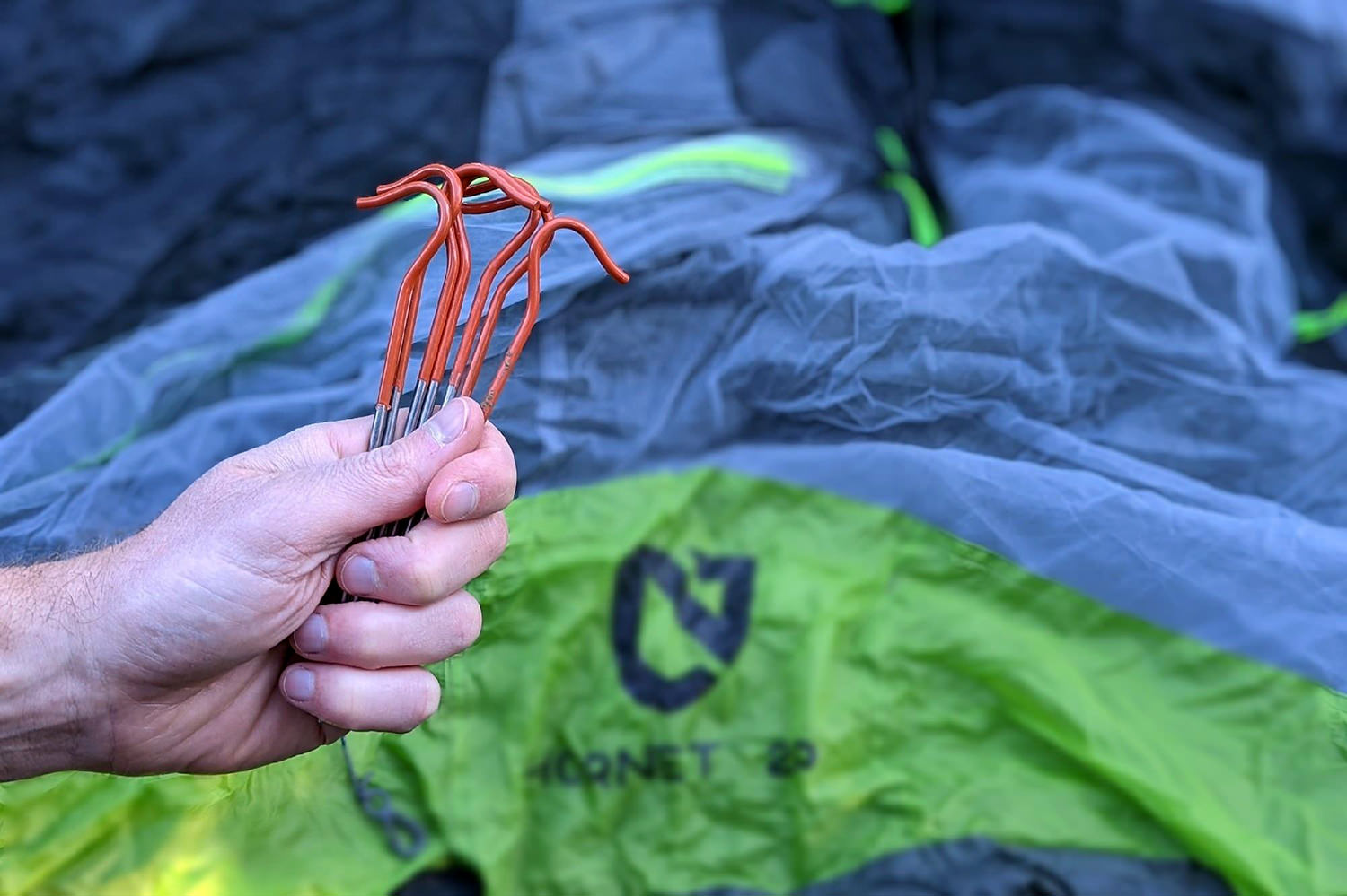 A backpacker's hand holding a set of Vargo Titanium Shepherds Hook tent stakes in his hand with a NEMO tent in the background