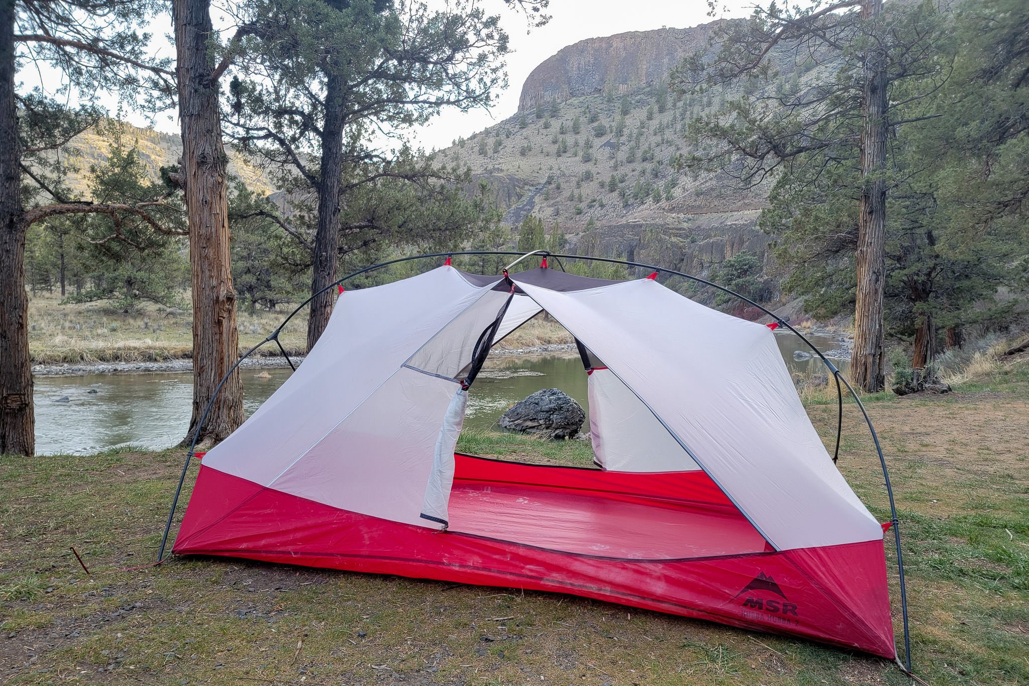 A white and red tent is setup next to a lake in the woods with a mountain in the background.