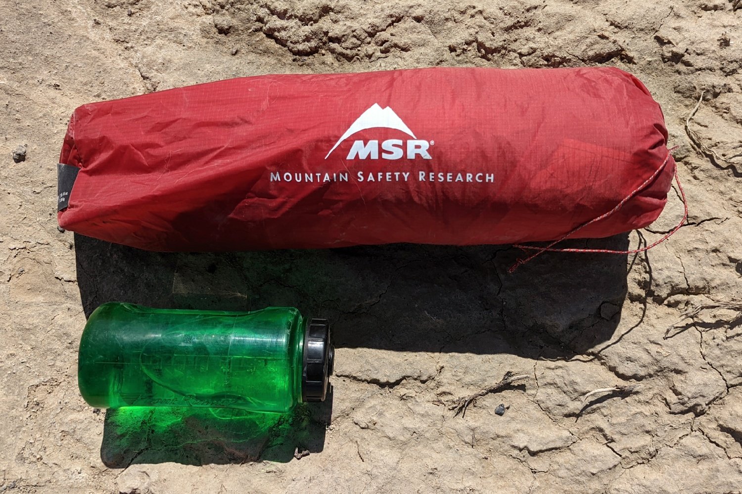 A tent fully packed into its stuff sack laying next a water bottle on the sandy ground.