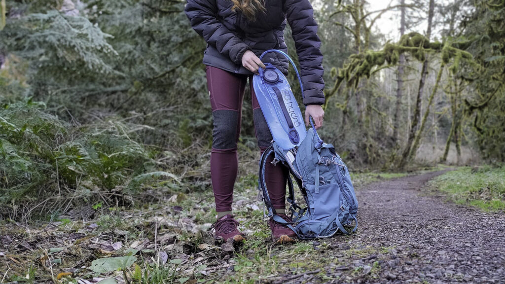 Waist-down view of a female hiker pulling the Gregory 3D Hydro hydration bladder out of her backpack along a trail