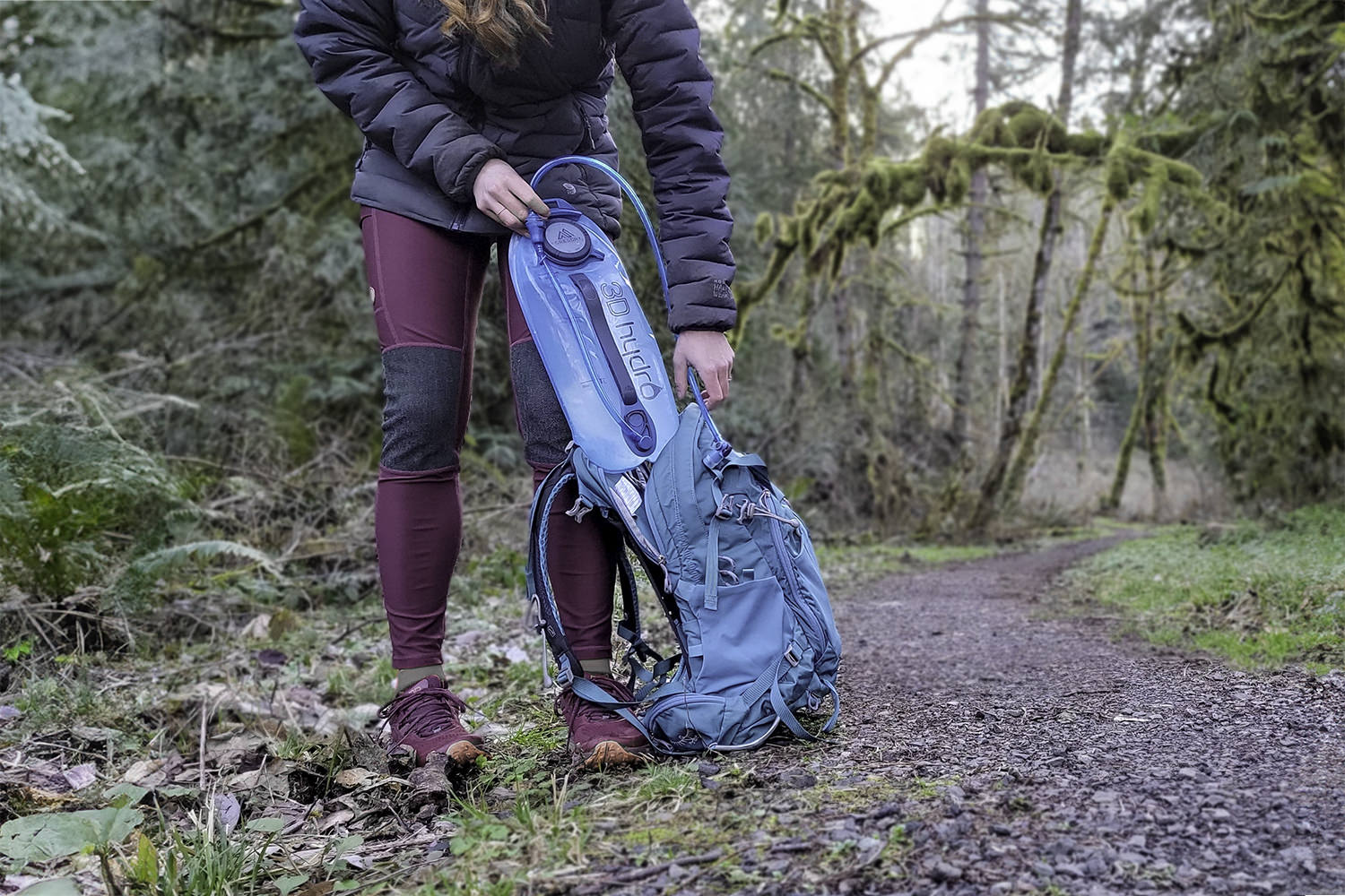 Waist-down view of a female hiker pulling the Gregory 3D Hydro hydration bladder out of her backpack along a trail