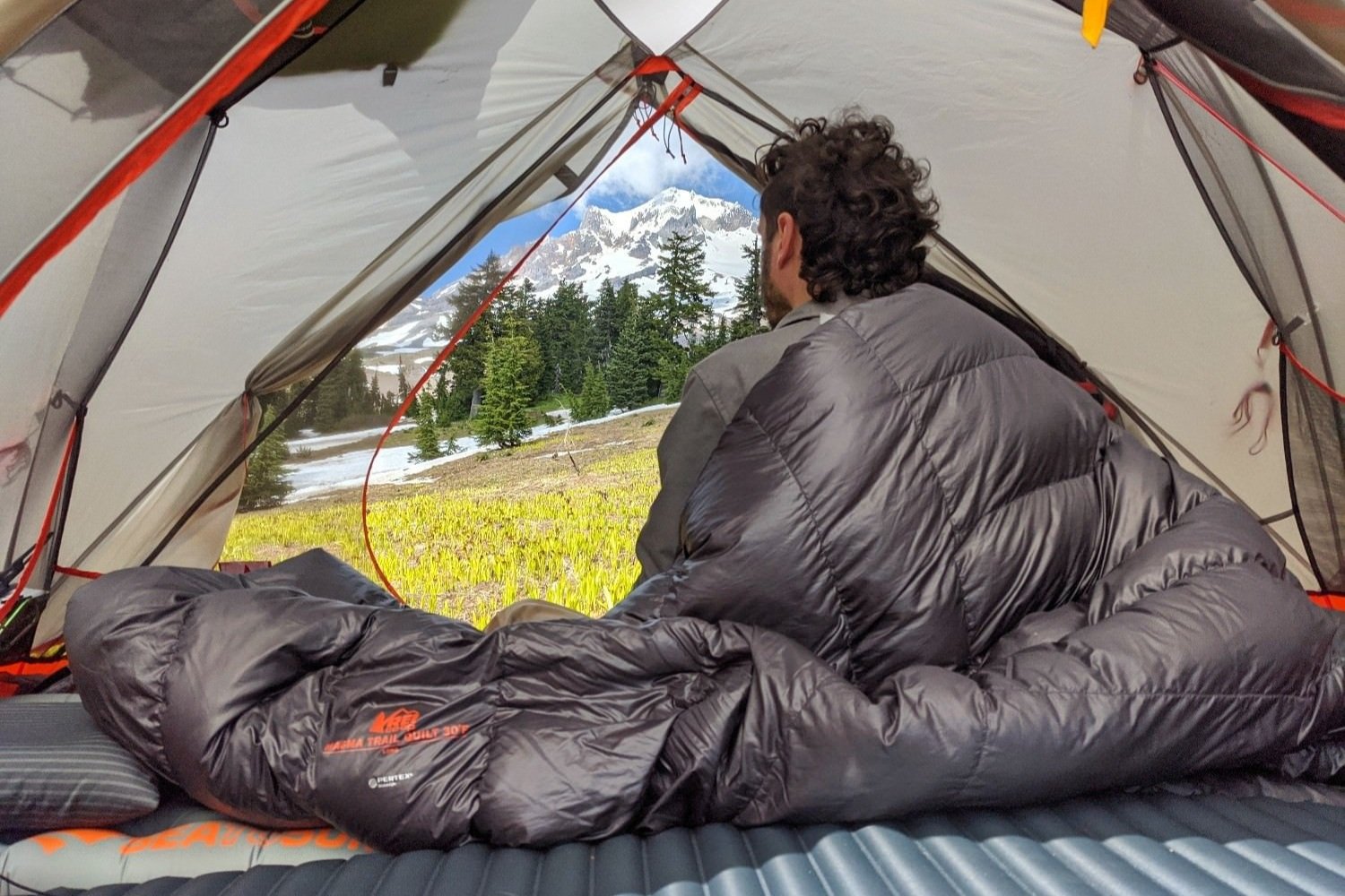 A hiker sitting in a tent wrapped in a Magma Trail Quilt in front of a mountain view