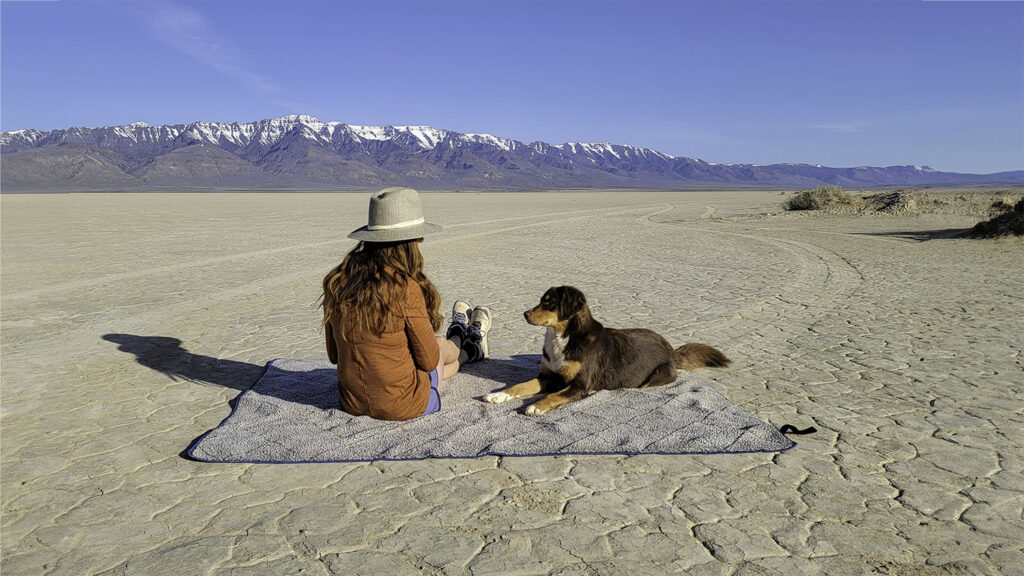 A woman and a dog sitting on the Oceas Sherpa Fleece camping blanket in the Alvord Desert with the Steens Mountains in the background