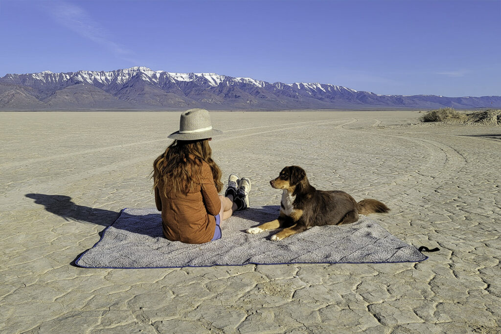 A woman and a dog sitting on the Oceas Sherpa Fleece camping blanket in the Alvord Desert with the Steens Mountains in the background