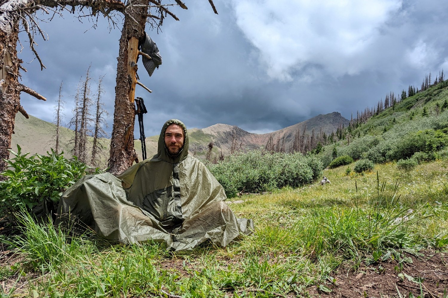 A hiker sitting under the Six Moon Designs Gatewood Cape among some trees while a storm forms on the Colorado Trail