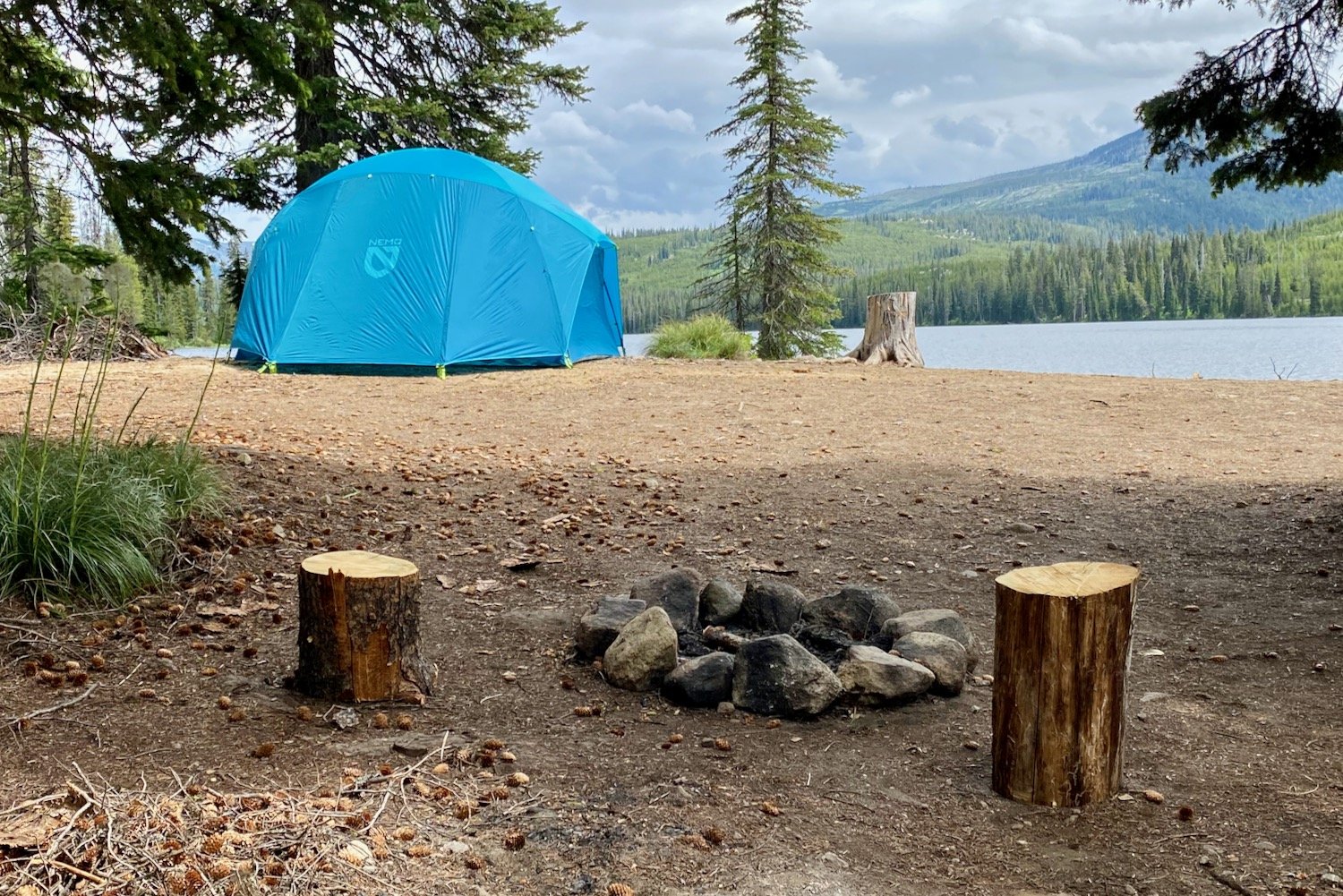 A campfire ring in the foreground with the NEMO Aurora Highrise Tent and a lake in the background.