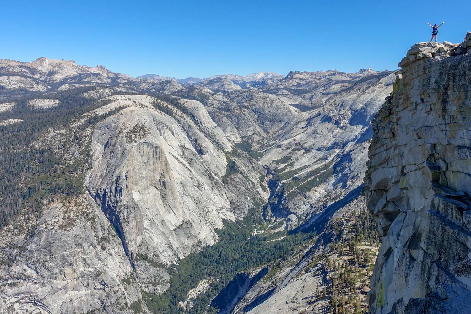 backpacker poses atop a granite wall with more granite rock walls lining a green valley below on a sunny day.