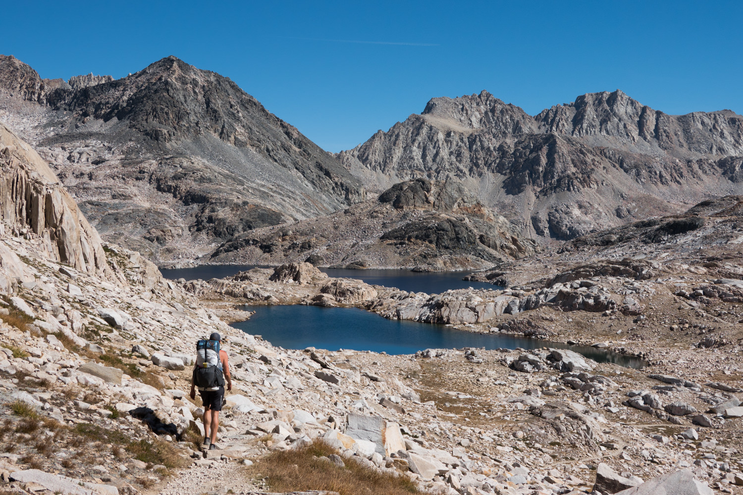 Backpacker walking toward two alpine lakes surrounded by rocky mountains on the John Muir Trail